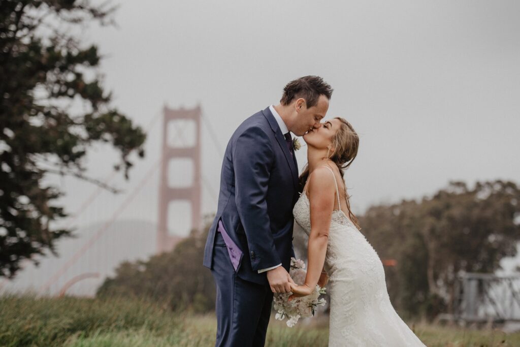 A bride and groom kissing in front of the golden gate bridge.