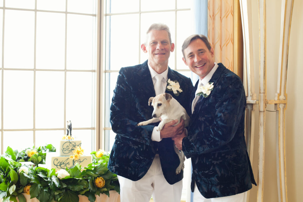 Two men holding a dog in front of a window.