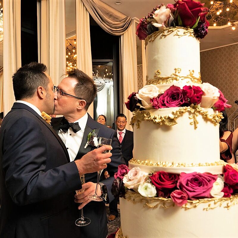Two men in suits are kissing next to a large cake.