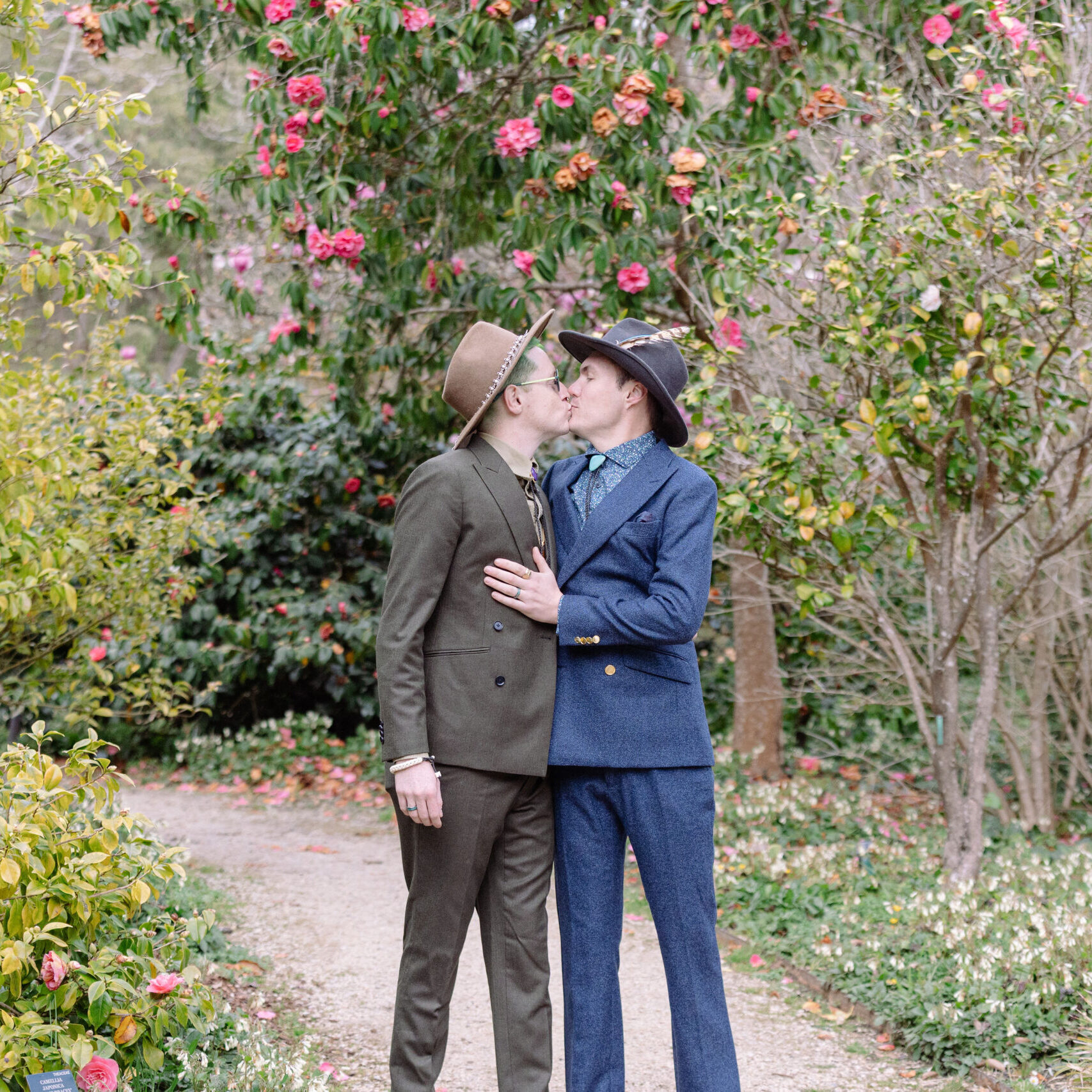 JV8A4590 Two men kissing in a garden with pink flowers.